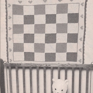 May include: A square crochet blanket featuring a checkerboard pattern of alternating light and dark squares. The border is decorated with small heart and leaf motifs, finished with a scalloped edge. The blanket is displayed above a wooden crib.