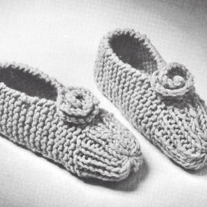 May include: A pair of light-coloured, hand-knitted slippers featuring a chunky yarn texture and a circular, rosette-style embellishment on the top of each foot. The slippers have a rounded toe design and a thick, ribbed knit construction throughout.