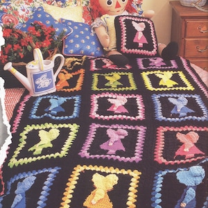 May include: A black crochet blanket featuring multiple square panels, each containing a colorful silhouette of a figure wearing a bonnet and dress. The squares are bordered in various bright colors including yellow, pink, blue, and green.