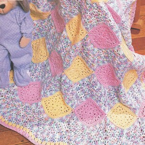 Peut inclure: Une couverture au crochet présentant un motif de carrés alternés jaune et rose unis, reliés par un fil multicolore dans des tons de bleu, violet et blanc. Une peluche portant une grenouillère violet clair est posée sur la couverture.