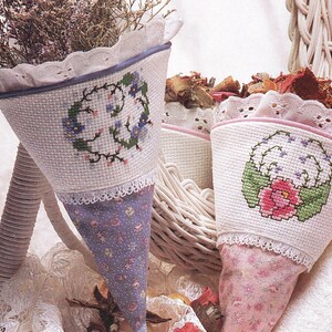 Peut inclure: Deux contenants en tissu en forme de cône avec des panneaux en point de croix blanc, broderies florales, bordures en dentelle et bases en tissu à motifs. Le cône gauche a un imprimé floral bleu, le droit un imprimé rose. Texte : Delicate and feminine floral cones are adorned with lace and pretty stitching. They'll hold flowers, potpourri or candy.