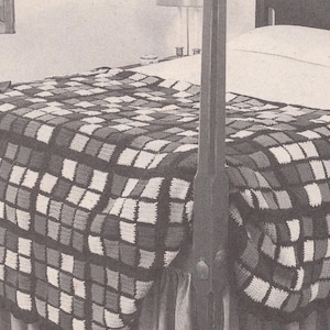 May include: A black and white photograph of a crocheted blanket featuring a repeating square grid pattern in alternating light and dark tones. The blanket is draped over the foot of a bed frame, with a doll positioned on the corner of the bed.