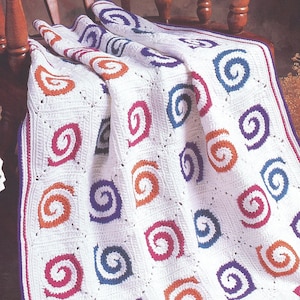 Peut inclure: Une couverture en crochet blanc composée de motifs carrés, chacun présentant une spirale centrale dans des tons orange, magenta et bleu. La couverture possède une bordure décorative avec de fines rayures violettes et magenta, en fil texturé.