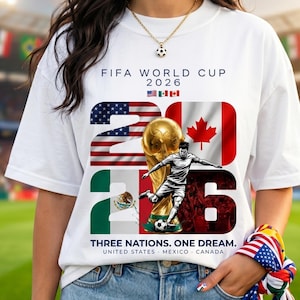 May include: A white t-shirt featuring the text FIFA WORLD CUP 2026, the flags of the USA, Mexico, and Canada, and a graphic of a soccer player kicking a ball next to a gold trophy. The text THREE NATIONS. ONE DREAM. UNITED STATES. MEXICO. CANADA is below.