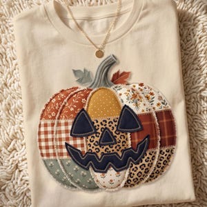 May include: A cream-colored t-shirt featuring a large, central pumpkin applique made of various fabric patches including floral, plaid, leopard print, and polka dot patterns. The pumpkin has a dark navy jack-o'-lantern face and a gold-tone necklace.