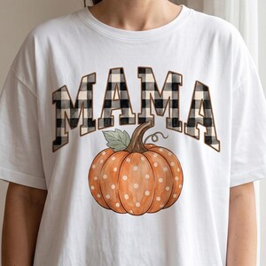 May include: A white short-sleeved t-shirt featuring the word MAMA in a black and white buffalo check pattern. Below the text is a graphic of an orange pumpkin with white polka dots, a brown stem, and a single green leaf, centred on the chest.