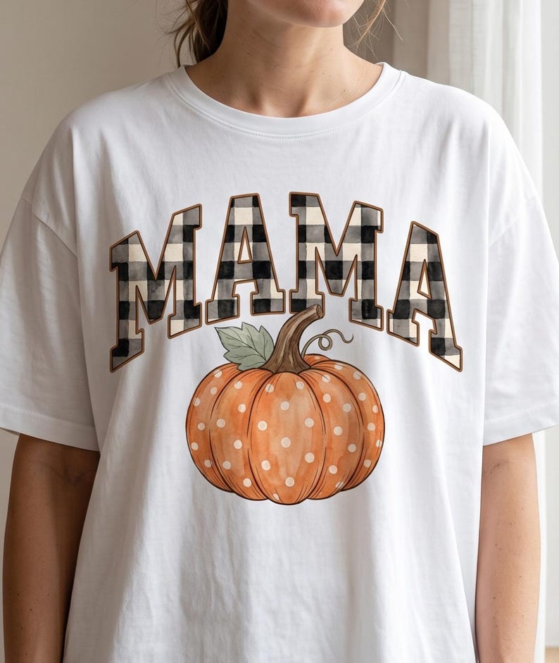 May include: A white short-sleeved t-shirt featuring the word MAMA in a black and white buffalo check pattern. Below the text is a graphic of an orange pumpkin with white polka dots, a brown stem, and a single green leaf, centred on the chest.