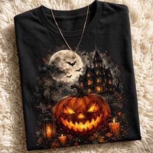May include: A black t-shirt featuring a graphic print of a glowing jack-o'-lantern, a haunted mansion, a full moon, and flying bats. The design includes lit lanterns and candles in a dark, atmospheric setting. A thin gold chain necklace rests on top.