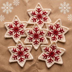 Puede incluir: Un conjunto de seis adornos de ganchillo en forma de estrella, cada uno con una base de color crema y un patrón de copo de nieve rojo vibrante bordado en el centro. Las estrellas de seis puntas están dispuestas en grupo.