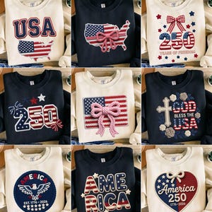 Op de afbeelding: Een verzameling crèmekleurige en marineblauwe sweatshirts met patriottische ontwerpen. De ontwerpen bevatten het woord "USA", de Amerikaanse vlag, het nummer "250" en het woord "America". Sommige ontwerpen hebben een strik of een kruis.