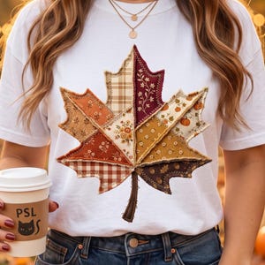 May include: A white t-shirt featuring a large, central maple leaf graphic composed of various fabric-style patches in autumn colours including orange, brown, and cream. The patches display patterns like plaid, floral, and pumpkins. A coffee cup with the text PSL is visible.