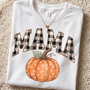 May include: A white short-sleeve t-shirt featuring the word MAMA in a black and white buffalo plaid pattern, positioned above a large orange pumpkin with white polka dots and a green leaf, accented by a gold-toned heart pendant necklace.