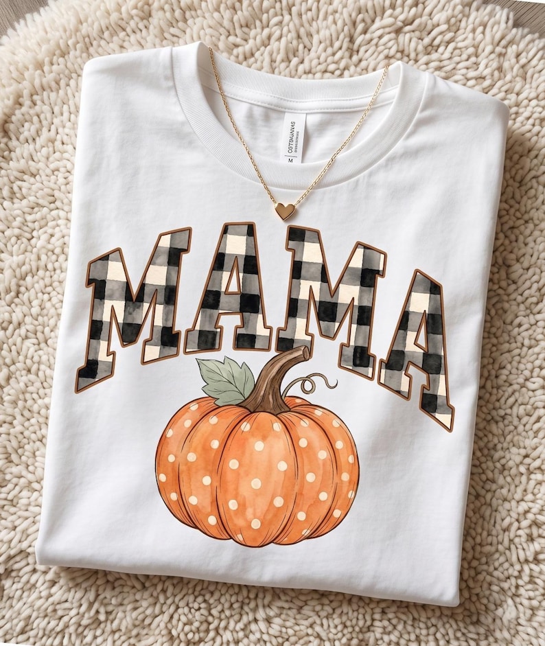 May include: A white short-sleeve t-shirt featuring the word MAMA in a black and white buffalo plaid pattern, positioned above a large orange pumpkin with white polka dots and a green leaf, accented by a gold-toned heart pendant necklace.