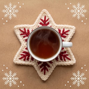 Puede incluir: Una taza de cerámica blanca llena de líquido marrón oscuro se sitúa centrada sobre un posavasos de ganchillo en forma de estrella. El posavasos tiene una base color crema con un patrón rojo cosido que recuerda a las agujas de pino.