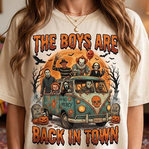 Può includere: Una t-shirt color crema con una stampa grafica di un furgone vintage pieno di personaggi dei film horror. Il testo recita THE BOYS ARE BACK IN TOWN, NO MERCY e SPOOKY1. Il design include zucche, lapidi e una luna piena sullo sfondo.