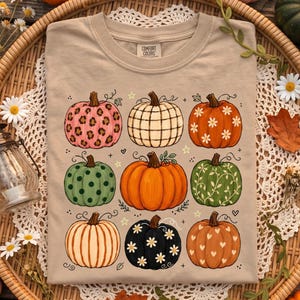 May include: A beige t-shirt featuring a graphic print of nine pumpkins in various patterns and colors, including leopard print, plaid, floral, polka dot, and heart designs. The shirt has a crew neckline and a Comfort Colors brand label.