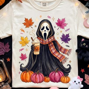 May include: A cream-colored short-sleeve t-shirt featuring a graphic of a ghost character wearing a plaid scarf and holding a pumpkin-themed coffee cup. The design includes colorful autumn leaves, small stars, and pumpkins in orange and pink hues.