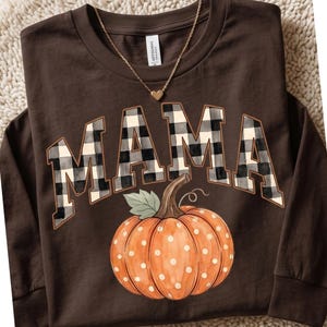 May include: A dark brown long-sleeve shirt featuring the word MAMA in a black and white buffalo plaid pattern. Below the text is a graphic of an orange pumpkin with white polka dots and a green leaf. A gold-tone heart pendant necklace is included.