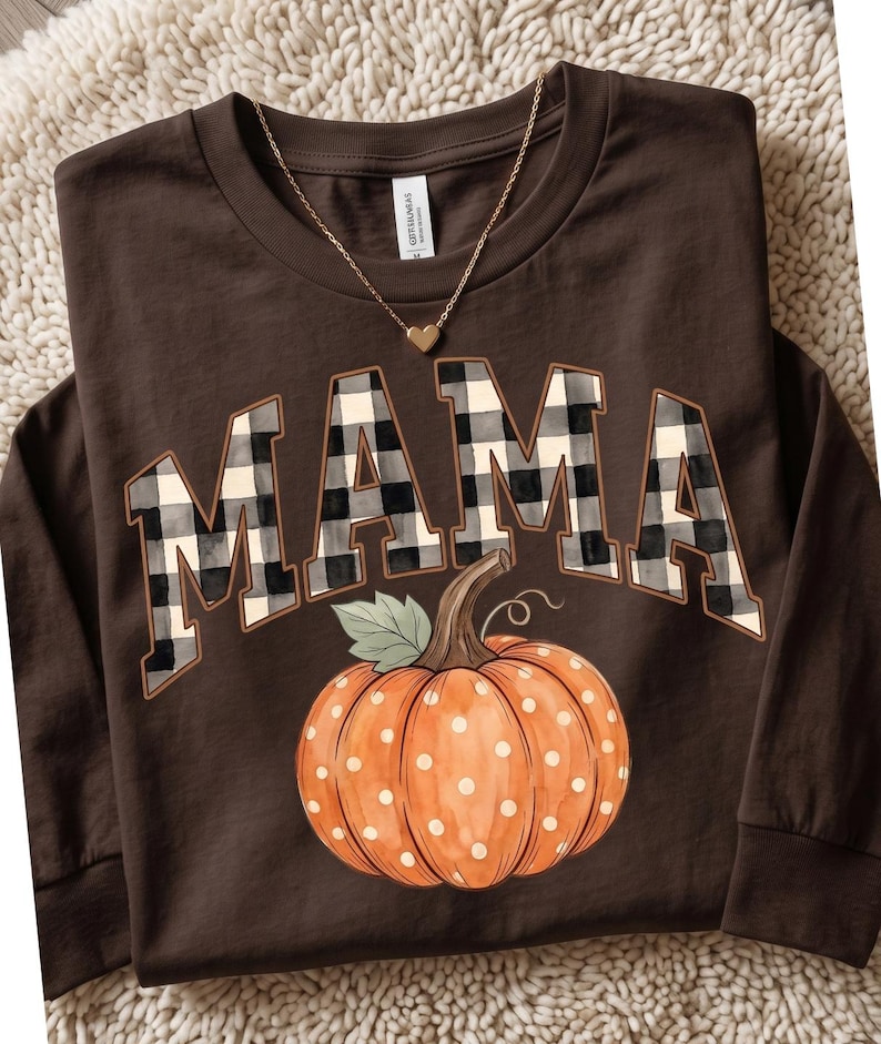 May include: A dark brown long-sleeve shirt featuring the word MAMA in a black and white buffalo plaid pattern. Below the text is a graphic of an orange pumpkin with white polka dots and a green leaf. A gold-tone heart pendant necklace is included.