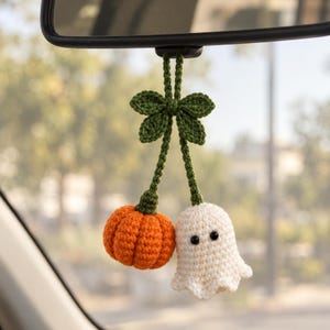 May include: A crochet car accessory hanging from a rearview mirror, featuring an orange pumpkin and a white ghost attached to green stems with a decorative green leaf bow at the top. The ghost has two small black bead eyes.