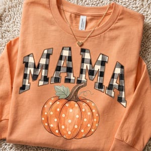 May include: An orange long-sleeve shirt featuring the word MAMA in a black and white buffalo plaid pattern. Below the text is a graphic of an orange pumpkin with white polka dots, a green leaf, and a brown stem. A gold heart necklace rests on the collar.