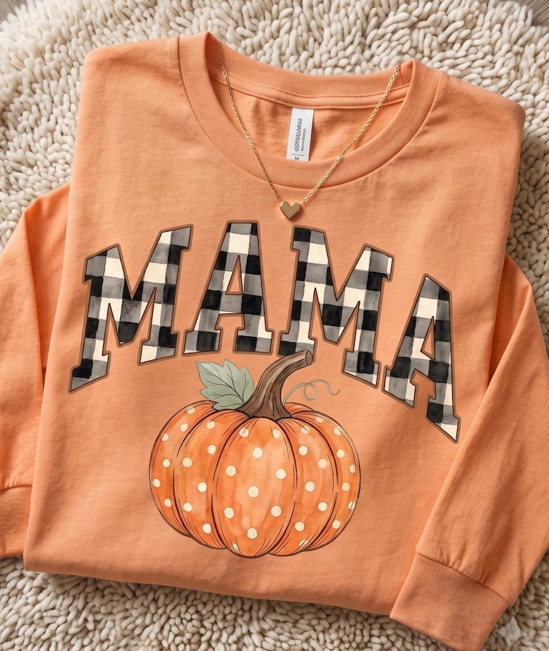 May include: An orange long-sleeve shirt featuring the word MAMA in a black and white buffalo plaid pattern. Below the text is a graphic of an orange pumpkin with white polka dots, a green leaf, and a brown stem. A gold heart necklace rests on the collar.