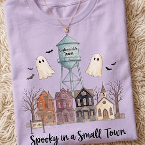 Puede incluir: Una camiseta de color morado claro con un gráfico de una torre de agua etiquetada como Customizable Town, dos fantasmas, murciélagos y una hilera de casas. Un letrero dice ZIP CODE. El texto Spooky in a Small Town está impreso abajo.