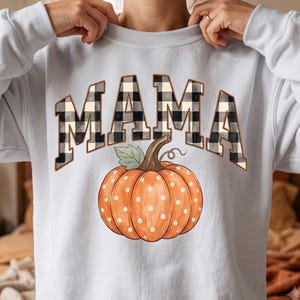May include: A light grey crewneck sweatshirt featuring the word MAMA in a black and white buffalo plaid pattern. Below the text is a large orange pumpkin with white polka dots, a brown stem, and a single green leaf, centred on the chest.