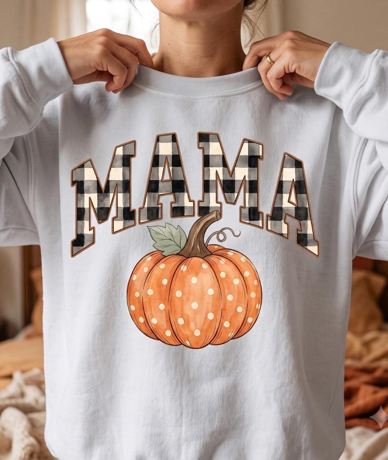 May include: A light grey crewneck sweatshirt featuring the word MAMA in a black and white buffalo plaid pattern. Below the text is a large orange pumpkin with white polka dots, a brown stem, and a single green leaf, centred on the chest.