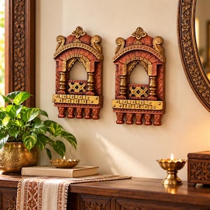 May include: Two rectangular wall hangings featuring an arched window design with intricate carvings. The pieces are finished in shades of terracotta red and metallic gold, showcasing detailed columns, a lattice railing, and small hanging finials.