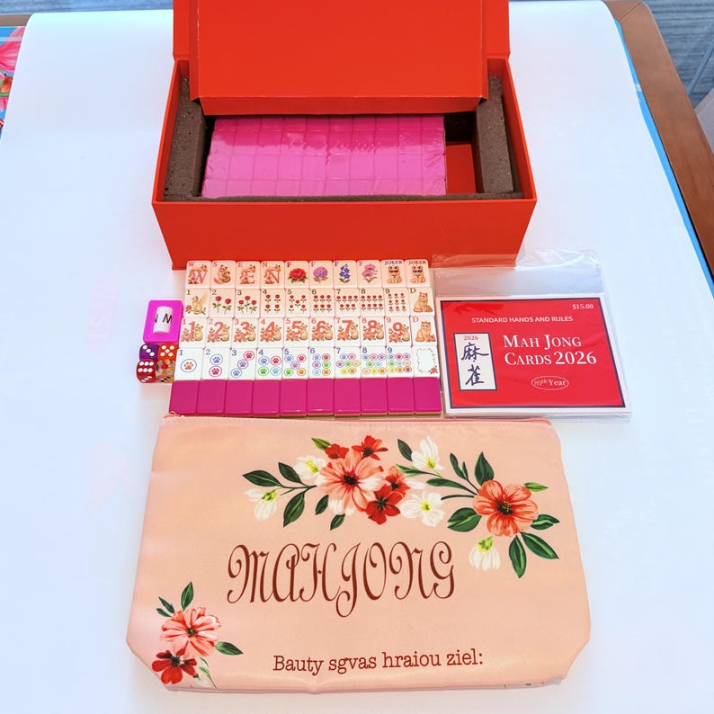 May include: A Mah Jong set featuring a bright orange storage box, a collection of white tiles with colorful floral and numerical designs, a set of dice, and a red booklet titled Mah Jong Cards 2026. A peach-colored zippered pouch displays floral art.