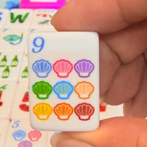 May include: A white rectangular mahjong tile featuring the blue number 9 in the top left corner and a grid of nine colorful seashell illustrations in shades of blue, pink, purple, green, light blue, orange, yellow, dark orange, and red.