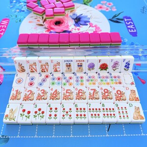 May include: A set of white mahjong tiles featuring colorful illustrations of cats, paw prints, flowers, and the letters N, W, E, F, and D. The tiles have pink and green backs and are arranged in rows on a blue mat with floral and directional text.