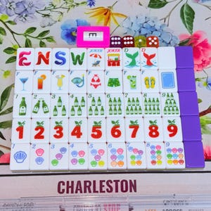 May include: A set of white mahjong tiles arranged in a grid, featuring colorful illustrations of letters, cocktails, beach items, bottles, strawberries with numbers, and seashells. A pink wind indicator and four small dice are placed on top.
