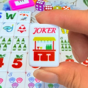 May include: A white rectangular mahjong tile featuring the red text JOKER above a graphic of a red bar counter with two stools, a yellow roof, and green bottles. The tile is held against a background of other tiles with various colorful icons.