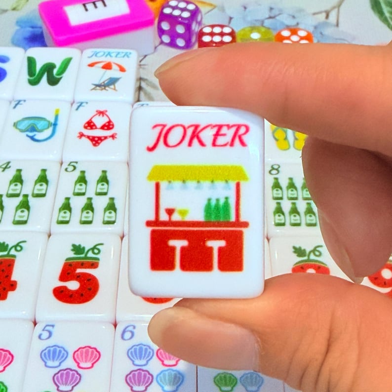May include: A white rectangular mahjong tile featuring the red text JOKER above a graphic of a red bar counter with two stools, a yellow roof, and green bottles. The tile is held against a background of other tiles with various colorful icons.