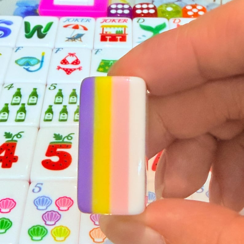 May include: A rectangular mahjong tile featuring vertical stripes of purple, yellow, light pink, and white. The tile is held upright against a background of other mahjong tiles decorated with various colorful icons including strawberries, shells, and bottles.