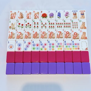 May include: A set of rectangular mahjong tiles featuring cat-themed illustrations, floral designs, numbers, and paw prints. The tiles are arranged in rows, with some displaying letters, joker labels, and blank spaces, alongside solid magenta and purple tiles.