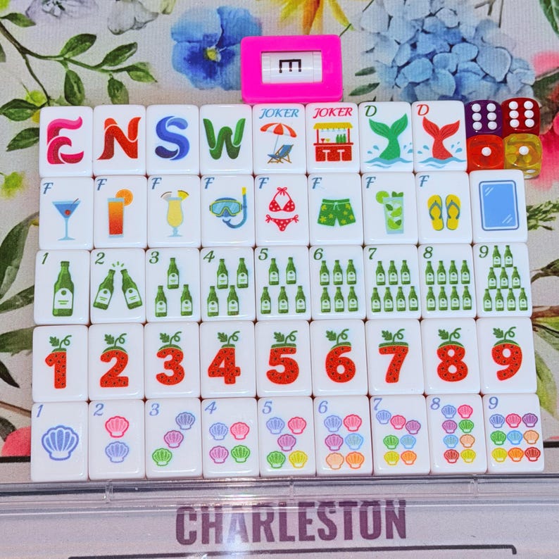 May include: A set of white mahjong tiles featuring colorful illustrations of cocktails, beach gear, strawberries, seashells, and green bottles, arranged in rows. A pink tile with the letter E and two translucent dice are included in the set.