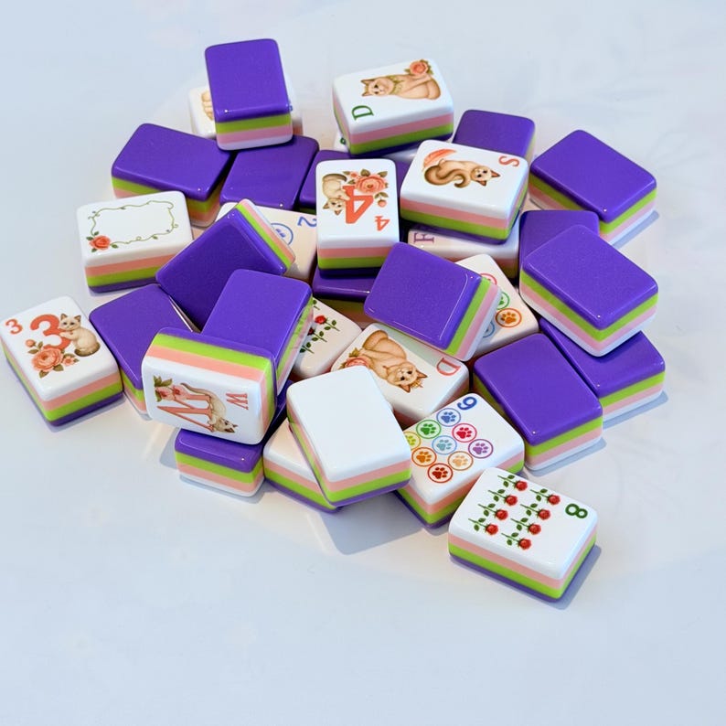 May include: A collection of rectangular mahjong tiles featuring a layered design with a purple top, a green middle, and a pink bottom. The tiles display various illustrations including cats, roses, paw prints, and numbers, with some tiles showing letters.