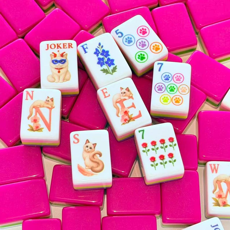 May include: A collection of mahjong tiles featuring bright pink backs and white fronts. The tiles display various illustrations including cats, flowers, paw prints, and roses, along with letters and numbers. Some tiles are labeled Joker, N, F, E, S, and W.
