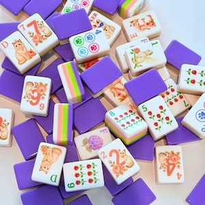 May include: A collection of rectangular mahjong tiles featuring purple backs and white fronts. The tiles display various illustrations including cats, roses, hydrangeas, and paw prints, alongside numbers and letters in orange, green, and blue ink.