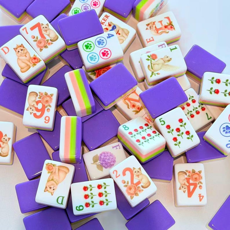 May include: A collection of rectangular mahjong tiles featuring purple backs and white fronts. The tiles display various illustrations including cats, roses, hydrangeas, and paw prints, alongside numbers and letters in orange, green, and blue ink.