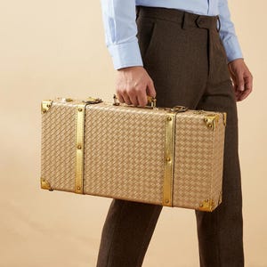 Peut inclure: Une valise rectangulaire au motif extérieur beige tressé, agrémentée de sangles métalliques verticales dorées et de renforts d'angle dorés. La valise comprend une poignée supérieure et des fermoirs métalliques sécurisés.