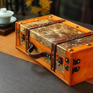 May include: A rectangular wooden storage chest with a light orange finish, featuring a gold-toned top panel with an embossed floral pattern. The chest includes dark brown leather-like straps, metal corner reinforcements, and a matching handle.