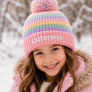 Puede incluir: Un gorro de punto con un patrón de rayas arcoíris en tonos pastel y un borde acanalado de color rosa sólido. La palabra Cuteness está bordada en blanco en la parte frontal del borde. El gorro tiene un pompón multicolor de lana a juego.