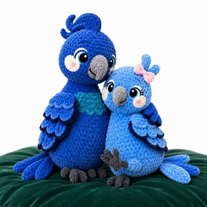 May include: Two crocheted parrot plush toys in shades of blue. The larger parrot is dark blue with a teal chest, while the smaller parrot is light blue with a pink bow on its head. Both have grey beaks, grey feet, and large embroidered eyes.