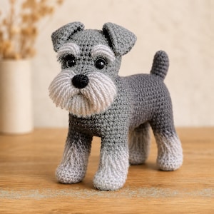 May include: A crocheted Schnauzer dog figurine featuring dark gray yarn on the body and head, with lighter gray yarn on the muzzle, eyebrows, and lower legs. The toy has black plastic eyes and a black yarn nose, standing on four legs with a short tail.