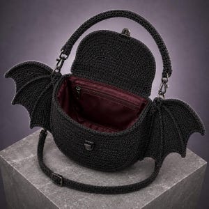 May include: A black crocheted handbag featuring two large bat-like wings on the sides, a top handle, and a detachable shoulder strap. The bag has a flap closure with a metal clasp and a deep burgundy interior lining with a zippered pocket inside.