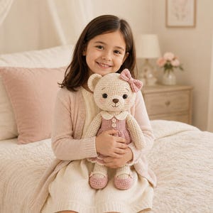 May include: A crocheted teddy bear toy featuring cream-colored yarn, a dusty pink dress with a white collar, two small buttons on the bodice, a matching pink bow on its ear, and pink shoes. The bear has embroidered facial features and rosy cheeks.